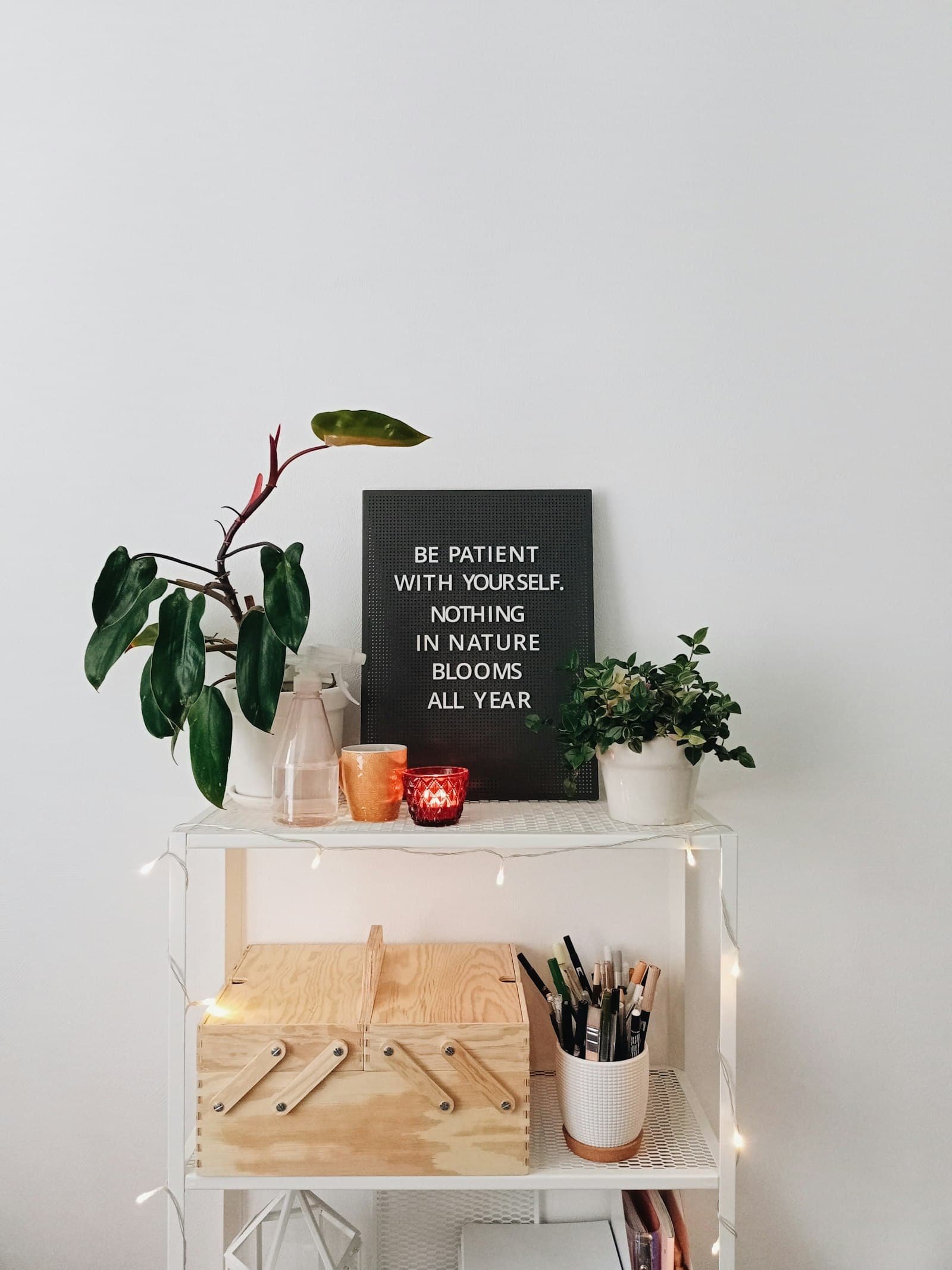 A white glass-shelved trolley against a pale wall: a trailing plant, small bottles, a lit candle and a framed card reading “be patient with yourself, nothing in nature blooms all year”